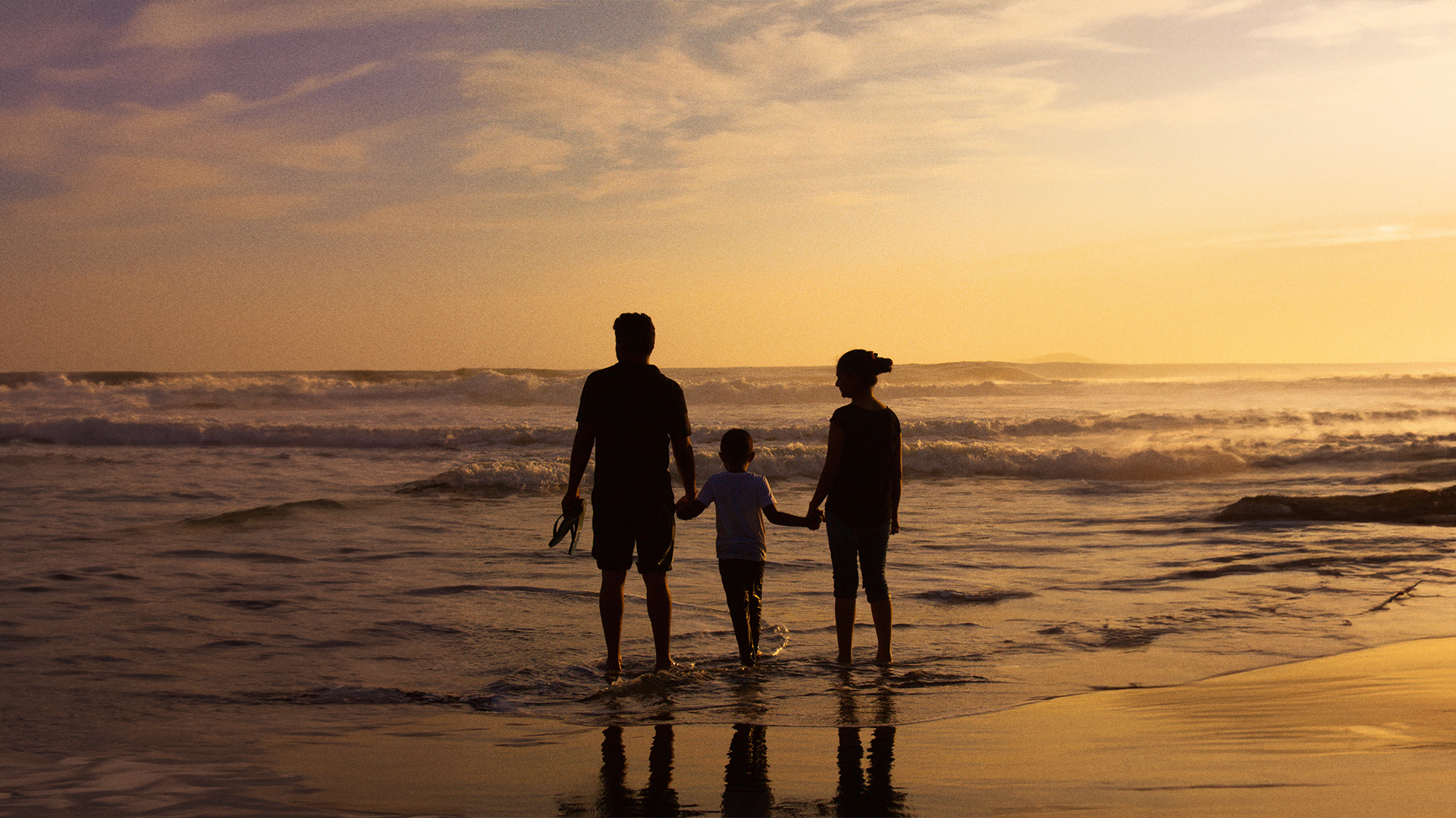 Familia de 3 agarrados de la mano, mirando el mar, en la playa al atardecer,